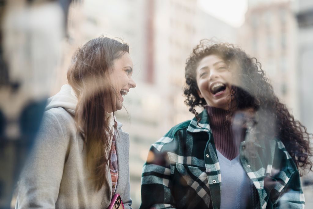 Through window of smiling multiracial female friends in outerwear having fun while standing near showcase on street on blurred background