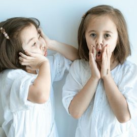Two young girls in white dresses express playful emotions indoors against a light background.