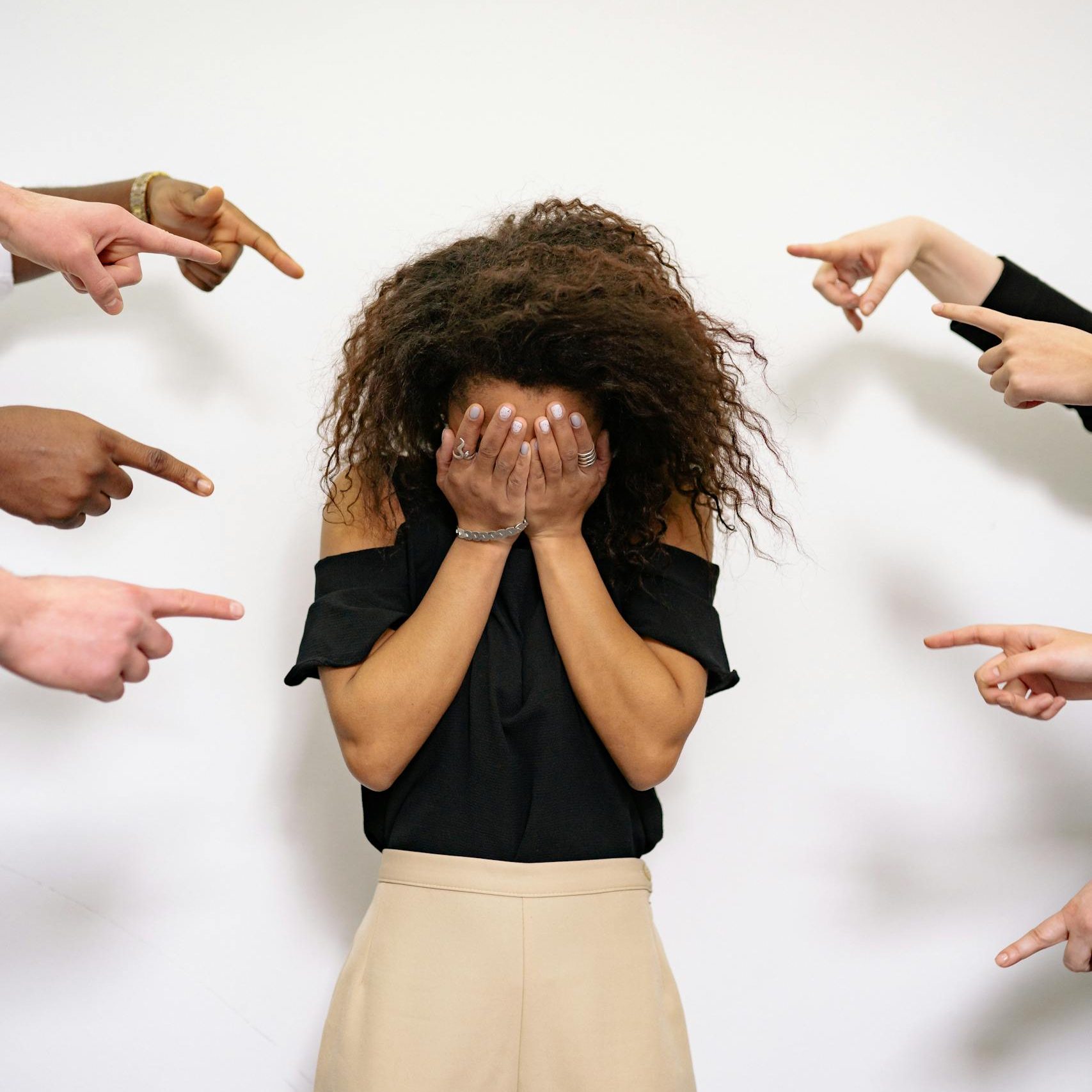 A stressed woman covers her face as multiple hands point accusatorily in a studio setting.