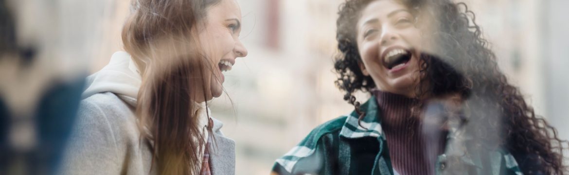 Through window of smiling multiracial female friends in outerwear having fun while standing near showcase on street on blurred background