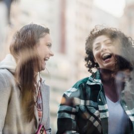 Through window of smiling multiracial female friends in outerwear having fun while standing near showcase on street on blurred background